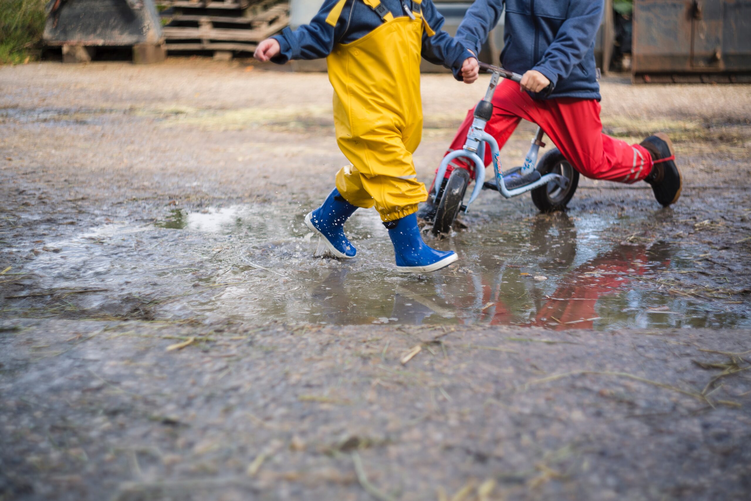 Two children play in white sand with a puddle. Their heads are not visible and one wears yellow rain overalls while the other sits on a bike wearing red rain trousers.